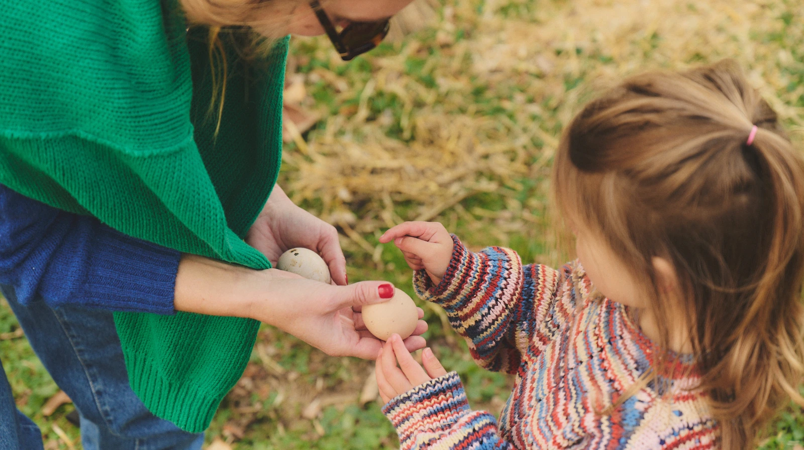 An adult hands fresh eggs to a child beside a backyard chicken coop.